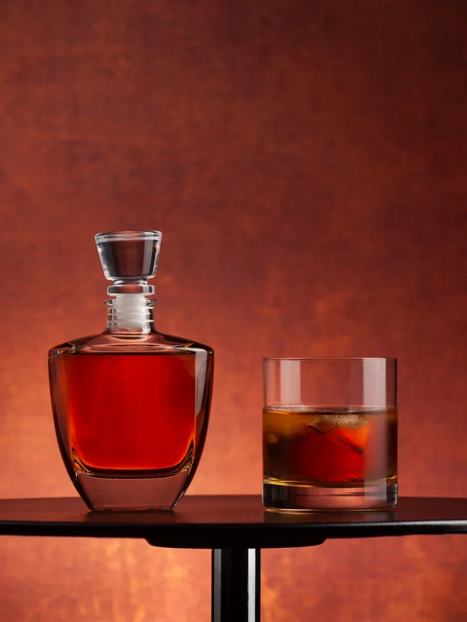 Unbranded heavy-base rectangular bottle with tapered sides and glass stopper, amber whisky, one tumbler against glowing rust background.