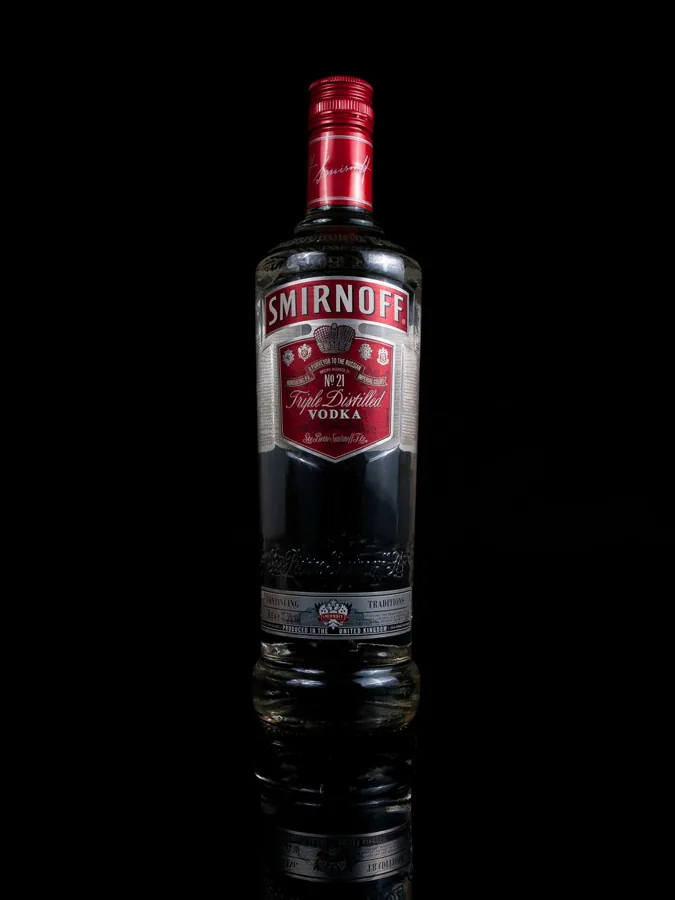 Single tall Smirnoff vodka bottle, long neck, screw cap and embossed lower body against black background with reflection.