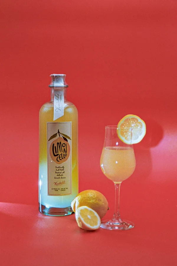 Tall limoncello bottle with heavy clear base, narrow neck and cork top on vivid coral backdrop with lemons and one glass.