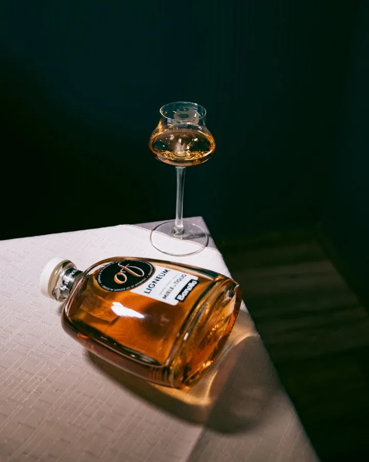 Low broad rectangular LIGNEUM grappa bottle laid diagonally on white table, with stemmed tasting glass against dark green.