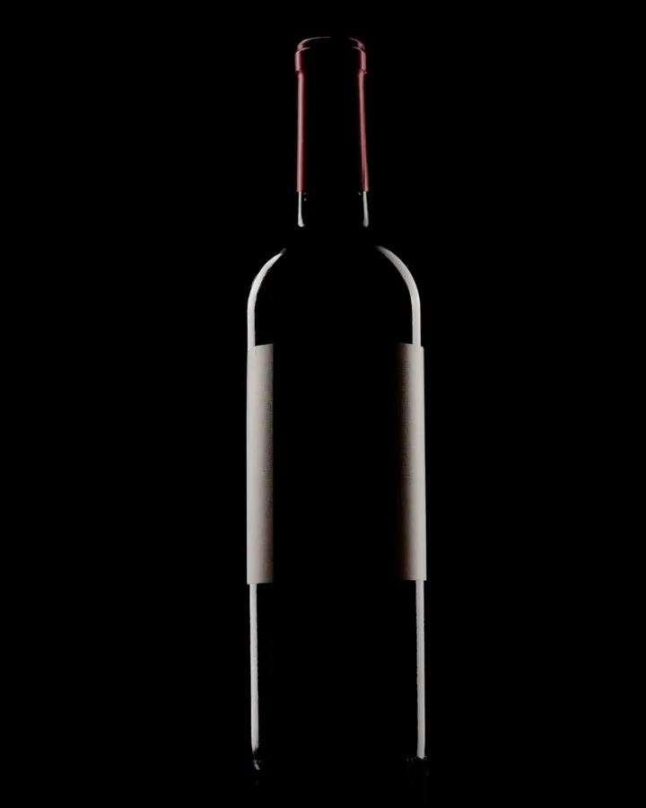 Full straight-shouldered wine bottle on black, red capsule, blank label and rim lighting. Strong portrait product image.