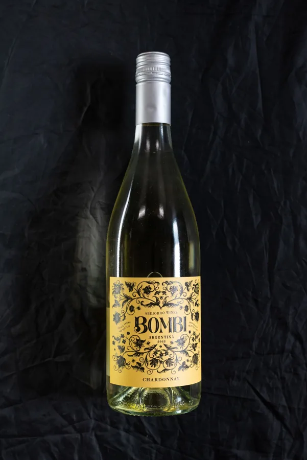 Complete sloping-shoulder clear Chardonnay bottle with silver screw cap and ornate yellow label on charcoal fabric. Good physical shape reference; Bombi brand readable. Shape category is a visual assessment, not geographic wine origin.