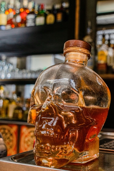 Clear skull-shaped glass spirits bottle with amber liquid and broad wooden stopper, photographed against a bar background.