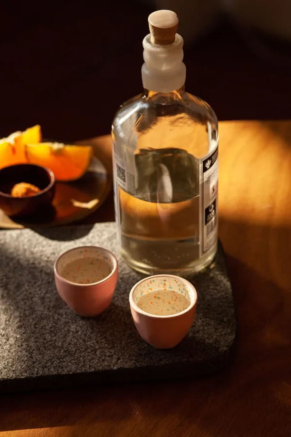Clear cylindrical bottle, rounded shoulders and white waxed neck with cork, lit by warm sun beside small ceramic cups and citrus.