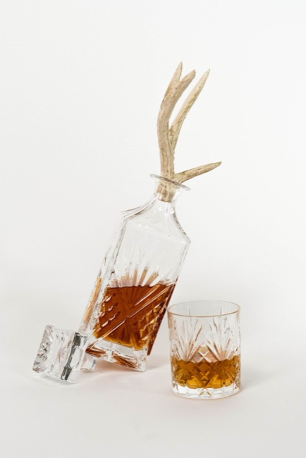 Angular clear cut-glass whisky decanter tilted on its base, with an antler-style sculptural stopper and a matching tumbler on a pale background.